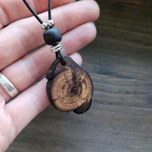 A wooden pendant necklace. The pendant is a dark brown, irregularly shaped piece of wood with visible grain and a natural edge. It hangs from a black leather cord, accented with a black bead and silver-coloured metal beads.