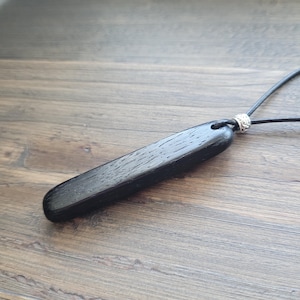 Op de afbeelding: Een zwarte houten hangerketting met een rechthoekige vorm. De hanger hangt aan een zwart koord en heeft een decoratieve zilveren kraal. De houtnerf is zichtbaar op de hanger.