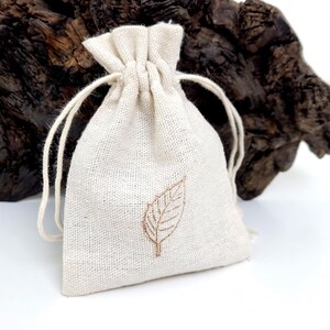 A sustainable packing bag for necklace. White linen jewellery drawstring pouch. The linen bag has brown leaf stamp in the middle and two white strings. The bag is displayed against the branch and white background, suggesting a sophisticated and eco-friendly jewellery packaging.
