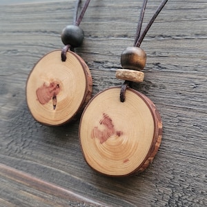 Pode incluir: Dois colares com pingentes de madeira. Cada pingente é uma fatia circular de madeira marrom clara com uma borda de casca marrom escura. O grão da madeira é visível. Os pingentes penduram de cordões de couro marrom, com uma conta de madeira escura e uma conta de madeira mais clara.