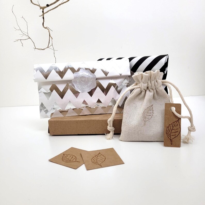 A collection of gift packaging, featuring a white and silver chevron-patterned envelope sealed with a clear wax stamp. A small drawstring bag and a tag, both with leaf designs, are also present. The packaging is on a white surface.