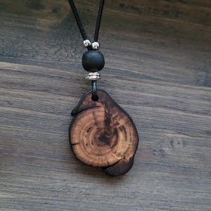 A wooden pendant necklace with a natural, organic shape. The dark brown pendant displays wood grain and a dark edge. It hangs from a black cord, with a black bead and silver-coloured accents.