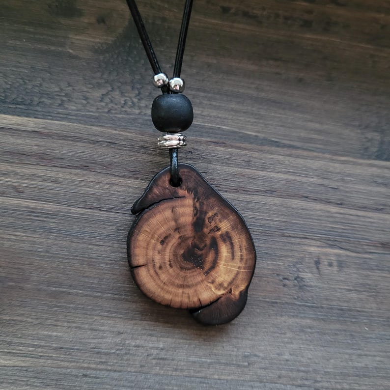 A wooden pendant necklace with a natural, organic shape. The dark brown pendant displays wood grain and a dark edge. It hangs from a black cord, with a black bead and silver-coloured accents.