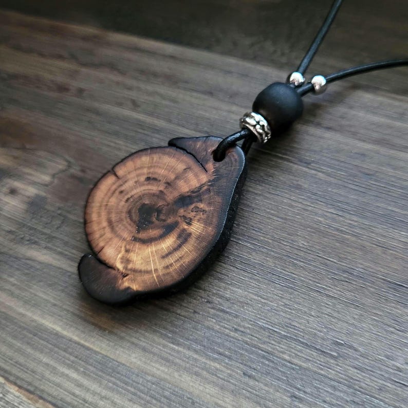 A natural wood woodland necklace charm. A wooden pendant necklace with a dark brown, organic-shaped wooden charm showing wood grain. The necklace has a black leather cord, a black bead, and silver-coloured accents. The pendant's shape is reminiscent of a cross-section of a tree trunk. A unique piece of wooden jewelry, with dark bohemian aesthetic. Handmade with care and attention.
