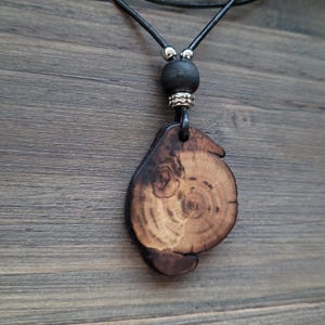 A dark woodland necklace with whimsy look. A wooden pendant necklace with a natural wood slice showing growth rings. The pendant is attached to a black leather cord, a black bead, and silver-toned accents. The wood has dark brown edges and a lighter brown centre. A unique piece of handcrafted bomenian jewelry with earthy aesthetic.