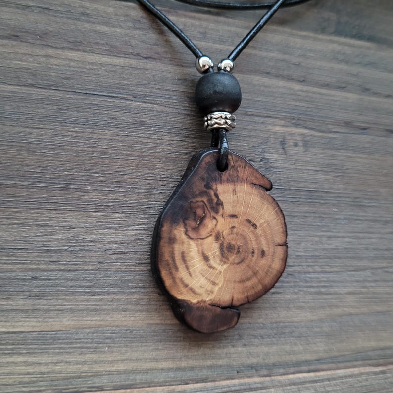 A dark woodland necklace with whimsy look. A wooden pendant necklace with a natural wood slice showing growth rings. The pendant is attached to a black leather cord, a black bead, and silver-toned accents. The wood has dark brown edges and a lighter brown centre. A unique piece of handcrafted bomenian jewelry with earthy aesthetic.