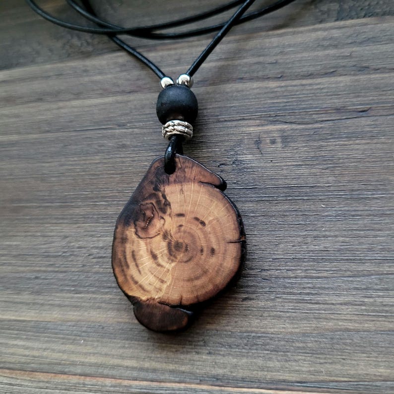 A wooden pendant necklace featuring a natural wood slice pendant with visible growth rings and a dark, textured edge. It hangs from a black leather cord, with a black bead and silver-toned accents. The necklace is displayed on a wooden surface. The necklace is a bohemian, earthy accessory, perfect for everyday wear.