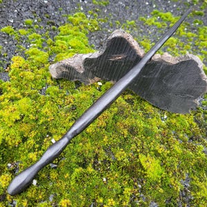 Pode incluir: Uma varinha escura, esculpida à mão, com uma ponta afiada, apoiada em um pedaço de madeira escura. A varinha está em um fundo de musgo verde vibrante e pedra escura e texturizada.
