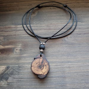 A handcrafted wooden pendant necklace. The pendant is a unique, organic shape with visible wood grain patterns in shades of brown and tan. The necklace features a black leather cord with an adjustable knot closure and a black bead.