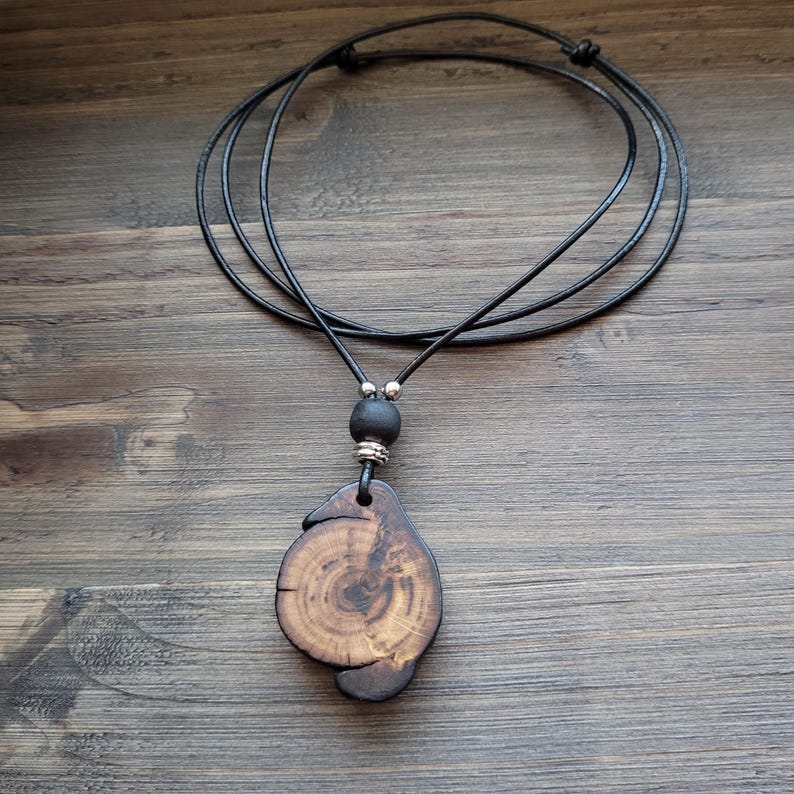 A handcrafted wooden pendant necklace. The pendant is a unique, organic shape with visible wood grain patterns in shades of brown and tan. The necklace features a black leather cord with an adjustable knot closure and a black bead.