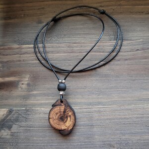 A handcrafted wooden pendant necklace. The pendant is a unique, organic shape with visible wood grain and a dark brown finish. The necklace features a black cord, a black bead, and silver-coloured accents. The pendant is approximately 4 cm in length.