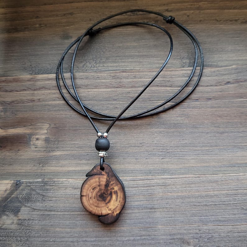 A handcrafted wooden pendant necklace. The pendant is a unique, organic shape with visible wood grain and a dark brown finish. The necklace features a black cord, a black bead, and silver-coloured accents. The pendant is approximately 4 cm in length.