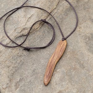 Handcrafted wooden pendant necklace of organic live edge oak wood in long irregular pin shape. The pendant is suspended on a dark brown cotton cord. An earthy vibe jewellery.