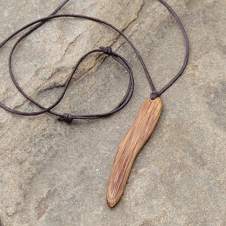 Handcrafted wooden pendant necklace of organic live edge oak wood in long irregular pin shape. The pendant is suspended on a dark brown cotton cord. An earthy vibe jewellery.