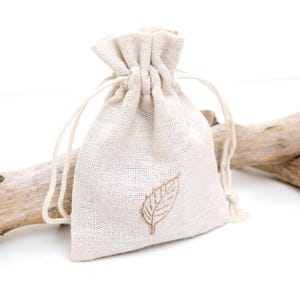 A sustainable packing bag for necklace. White linen jewellery drawstring pouch. The linen bag has brown leaf stamp in the middle and two white strings. The bag is displayed against the branch and white background, suggesting a sophisticated and eco-friendly jewellery packaging.