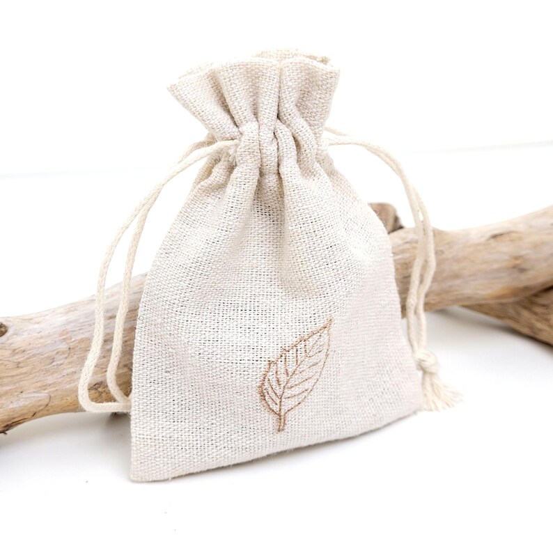 A sustainable packing bag for necklace. White linen jewellery drawstring pouch. The linen bag has brown leaf stamp in the middle and two white strings. The bag is displayed against the branch and white background, suggesting a sophisticated and eco-friendly jewellery packaging.