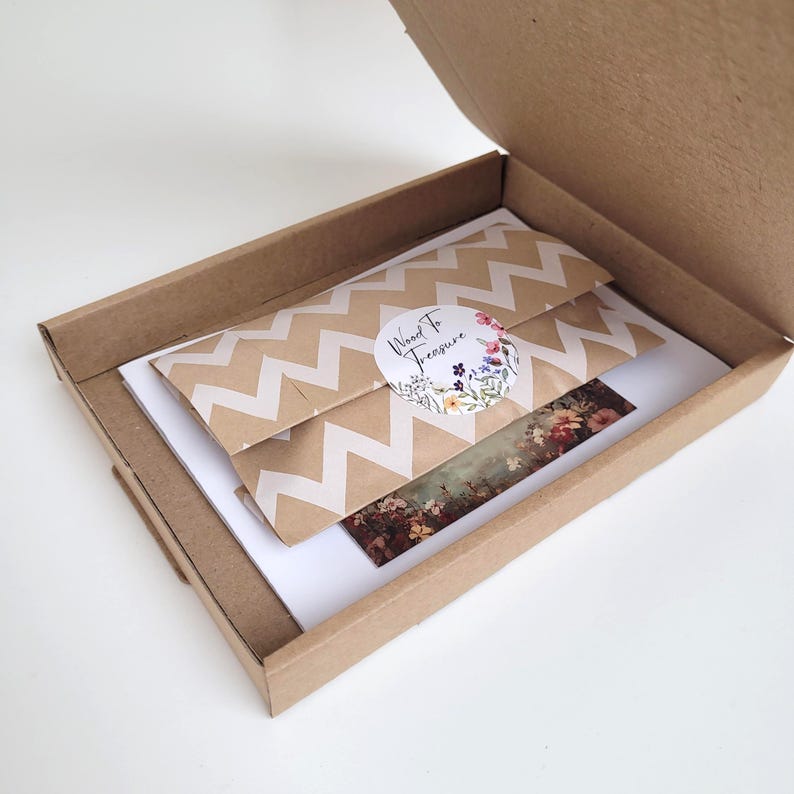 A brown cardboard box, open to reveal two packages with a white zigzag pattern. A floral sticker and a floral print are also visible. The sticker says "Wood To Treasure". The box is ready for posting.