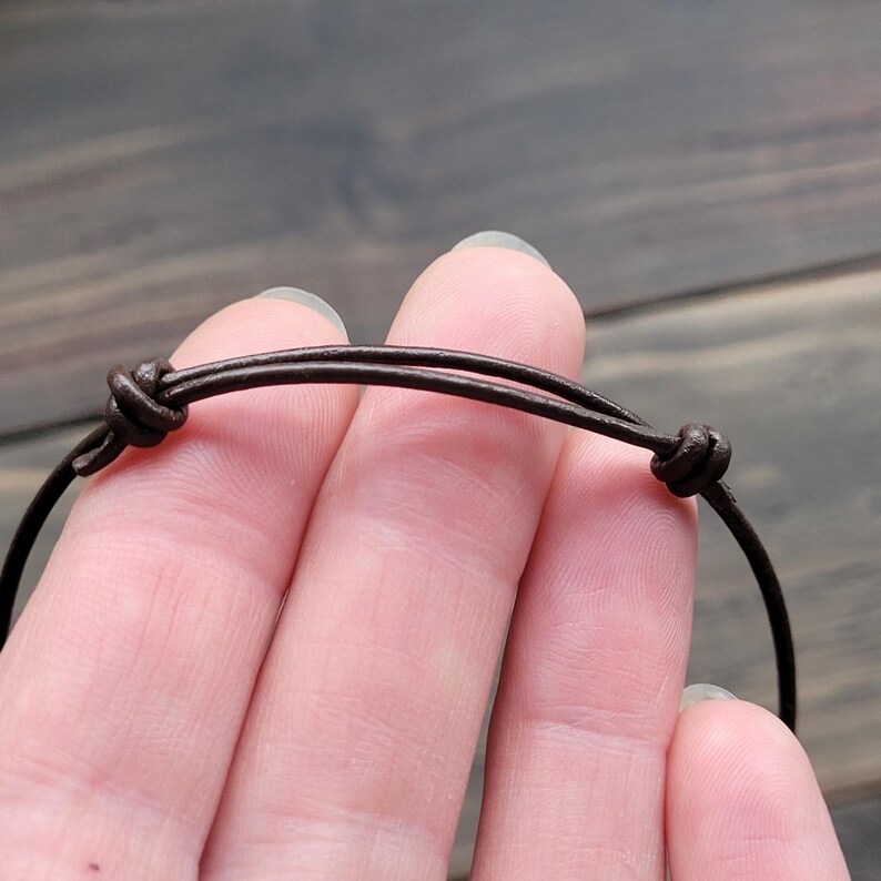 Close-up of a brown leather necklace with adjustable knots. The necklace is made of two thin, dark brown leather cords, knotted at each end for adjustability. The background is a blurred wooden surface.
