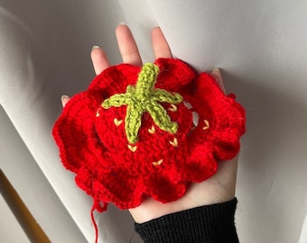 May include: A crocheted strawberry with a green star-shaped top and red body. The strawberry is held in a hand.