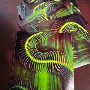 Op de afbeelding: Een stapel stickers met een close-up van een groen en zwart iriserend patroon. Het patroon lijkt op een opgerolde slang of een reeks overlappende ringen.