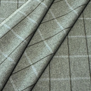 Op de afbeelding: Close-up van een gevouwen stof met een ruitpatroon. De stof is een gedempte groene kleur met een raster van donkerbruine en lichtblauwe lijnen. De stof heeft een getextureerde uitstraling.