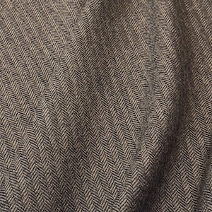 May include: Close-up of a folded fabric with a herringbone pattern. The fabric is a blend of brown and gray tones, creating a textured appearance. The diagonal lines of the weave are clearly visible, adding depth to the material.