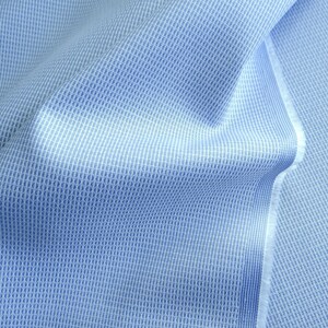 May include: Close-up of a light blue fabric with a textured, woven pattern. The fabric has a subtle grid-like design, with a lighter shade of blue creating a contrast. The edges of the fabric are visible, showing the raw, unfinished edge.