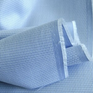 May include: Close-up of a light blue fabric with a textured weave. The fabric has a grid-like pattern with vertical stripes and a subtle, raised texture. The edges of the fabric are frayed, revealing the raw, unfinished edges of the material.