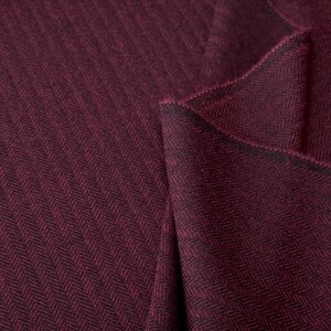 Könnte beinhalten: Nahaufnahme eines bordeauxfarbenen Stoffes mit Fischgrätmuster. Der Stoff ist gefaltet, wodurch die Textur und das Gewebe sichtbar werden. Die Farbe ist ein tiefes Rot mit dunklen Akzenten, geeignet für Kleidung oder Heimdekoration.