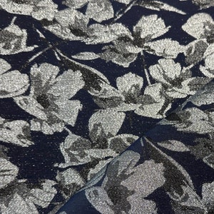 Könnte beinhalten: Nahaufnahme eines marineblauen Stoffes mit silbernem Blumenmuster. Die Blüten und Blätter glänzen metallisch. Der Stoff ist gefaltet, wodurch die Textur sichtbar wird. Geeignet für Kleidung oder Inneneinrichtung.