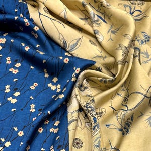 Könnte beinhalten: Ein gefalteter Seidenstoff mit zwei unterschiedlichen Mustern. Ein Abschnitt ist tiefblau mit weißen floralen Akzenten, während der andere hellgelb mit einem zarten schwarz-blauen botanischen Design mit Schmetterlingen und Blumen ist. Der Stoff hat eine weiche, fließende Textur.