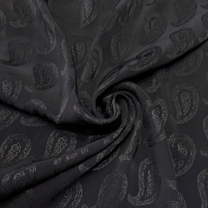 Könnte beinhalten: Schwarzer Stoff mit Paisley-Muster. Der Stoff ist einfarbig dunkel mit einem sich wiederholenden Design aus tropfenförmigen Paisley-Motiven. Der Stoff ist drapiert und gewirbelt, wodurch seine Textur sichtbar wird.