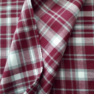 Può includere: Primo piano di un tessuto piegato con un motivo a quadri. Il tessuto presenta una combinazione di colori bordeaux, bianco e grigio. Il motivo a quadri è composto da linee intersecanti che creano un motivo a griglia. I bordi del tessuto sono visibili.