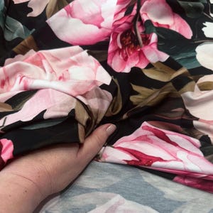 Pode incluir: Close-up de um tecido com um grande padr&atilde;o floral. O design apresenta grandes flores rosa e brancas com folhas verdes sobre um fundo preto. O tecido est&aacute; sendo segurado, mostrando sua textura e caimento.