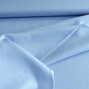 May include: Close-up of light blue fabric with a subtle grid pattern. The fabric is folded and draped, revealing the texture and weave. The color is a soft, pale blue, suitable for clothing or home decor projects.