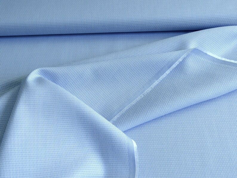 May include: Close-up of light blue fabric with a subtle grid pattern. The fabric is folded and draped, revealing the texture and weave. The color is a soft, pale blue, suitable for clothing or home decor projects.