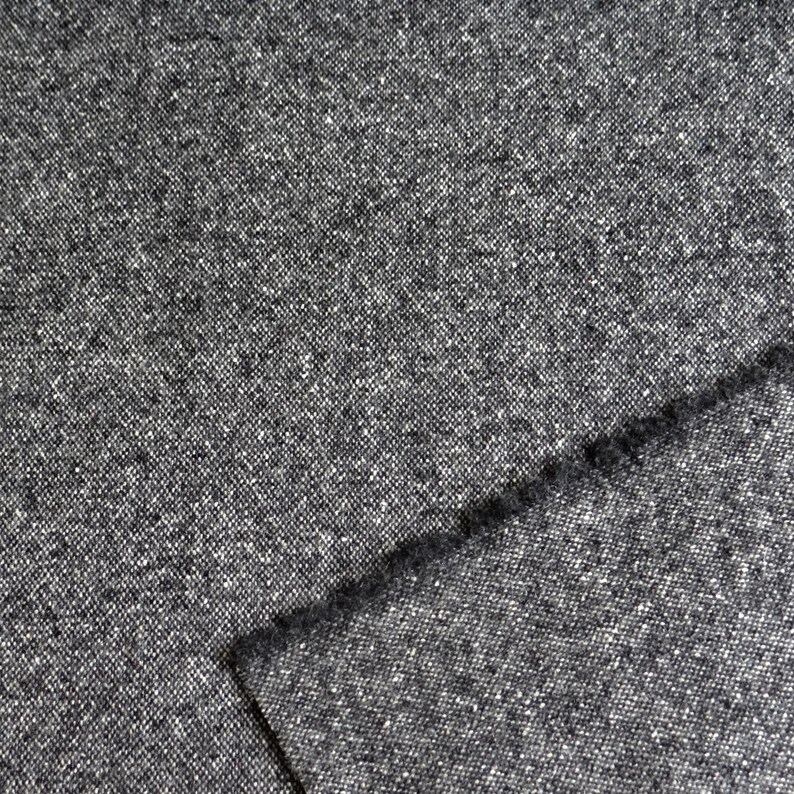 May include: Close-up of a gray tweed fabric with a textured surface. The fabric has a speckled appearance with small white and black flecks throughout. A folded edge reveals the fabric's thickness and weave.