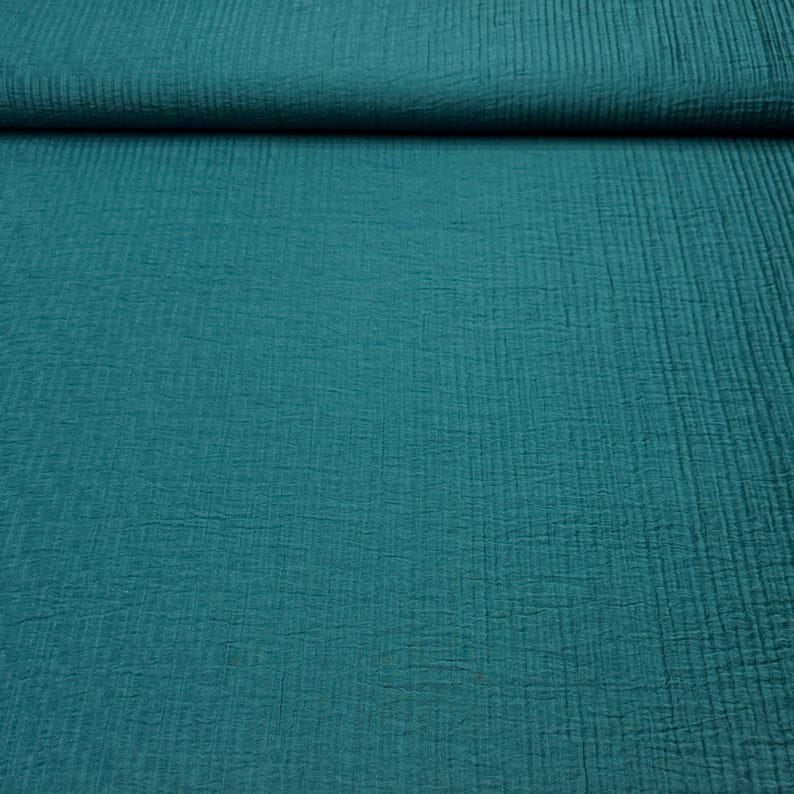 May include: Teal-colored fabric with a textured, crinkled appearance. The material is folded over at the top, revealing the layered texture. The fabric has a soft, matte finish and a subtle vertical pattern. Ideal for sewing projects.