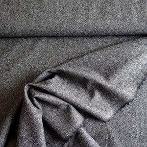 May include: A close-up shot of a folded piece of gray fabric. The fabric has a textured appearance and a subtle, speckled pattern. The folds create shadows and depth, highlighting the material's drape and texture. The edges of the fabric are visible.