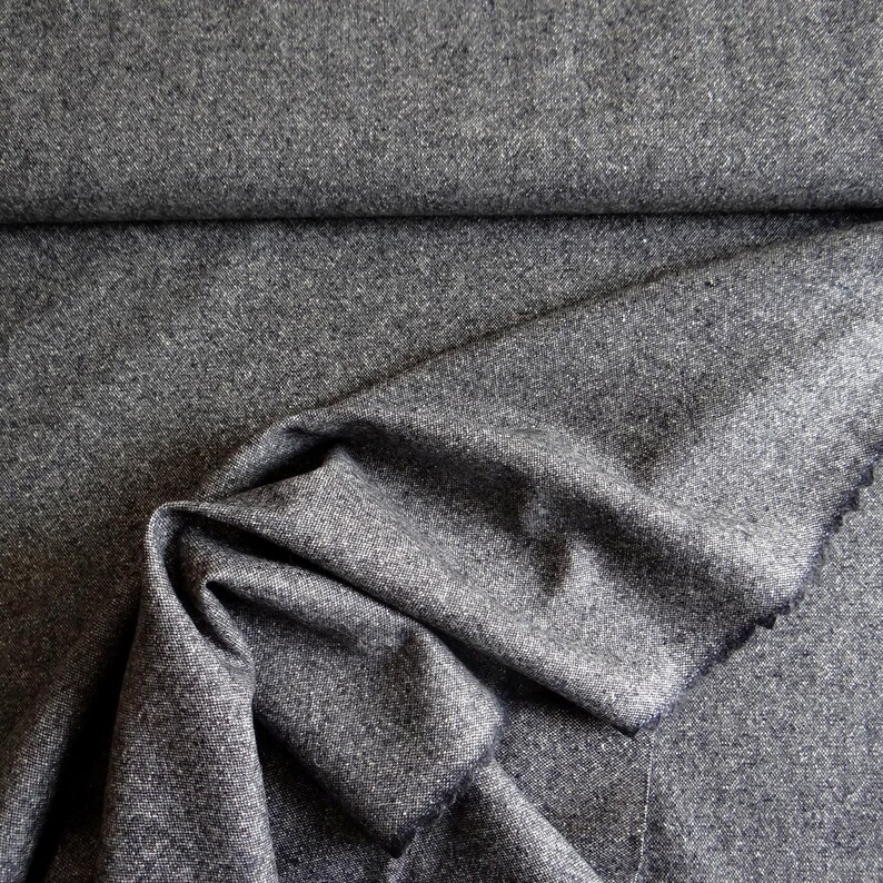 May include: A close-up shot of a folded piece of gray fabric. The fabric has a textured appearance and a subtle, speckled pattern. The folds create shadows and depth, highlighting the material's drape and texture. The edges of the fabric are visible.