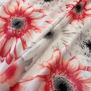 Könnte beinhalten: Nahaufnahme eines Stoffes mit floralem Muster. Das Design zeigt große Gerbera-Gänseblümchen in Rot-, Weiß- und Grautönen. Der Stoff ist gefaltet und zeigt die Textur und den geschichteten Effekt des Drucks.