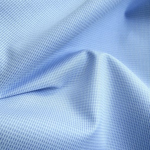 May include: Close-up of light blue fabric with a subtle grid pattern. The fabric is folded, creating soft shadows and highlighting the texture. The weave appears to be a fine, textured weave, suitable for shirts or other apparel.
