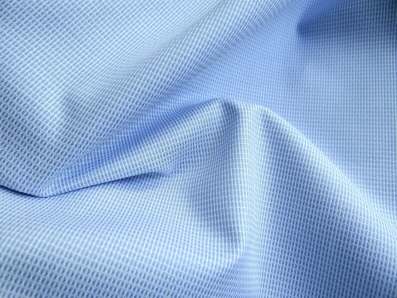 May include: Close-up of light blue fabric with a subtle grid pattern. The fabric is folded, creating soft shadows and highlighting the texture. The weave appears to be a fine, textured weave, suitable for shirts or other apparel.