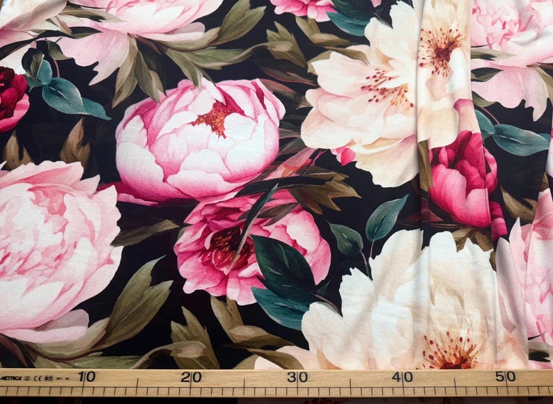 Pode incluir: Tecido floral com grandes pe&oacute;nias rosa e brancas sobre um fundo escuro. O tecido apresenta folhas verdes e uma r&eacute;gua na parte inferior, com medidas em cent&iacute;metros.