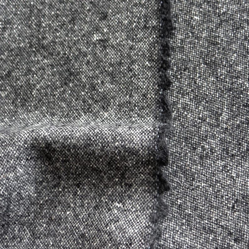 May include: Close-up of a textured fabric in shades of gray and white. The material has a woven appearance with a slightly rough texture. The edge of the fabric is visible, showing a raw, unfinished edge.