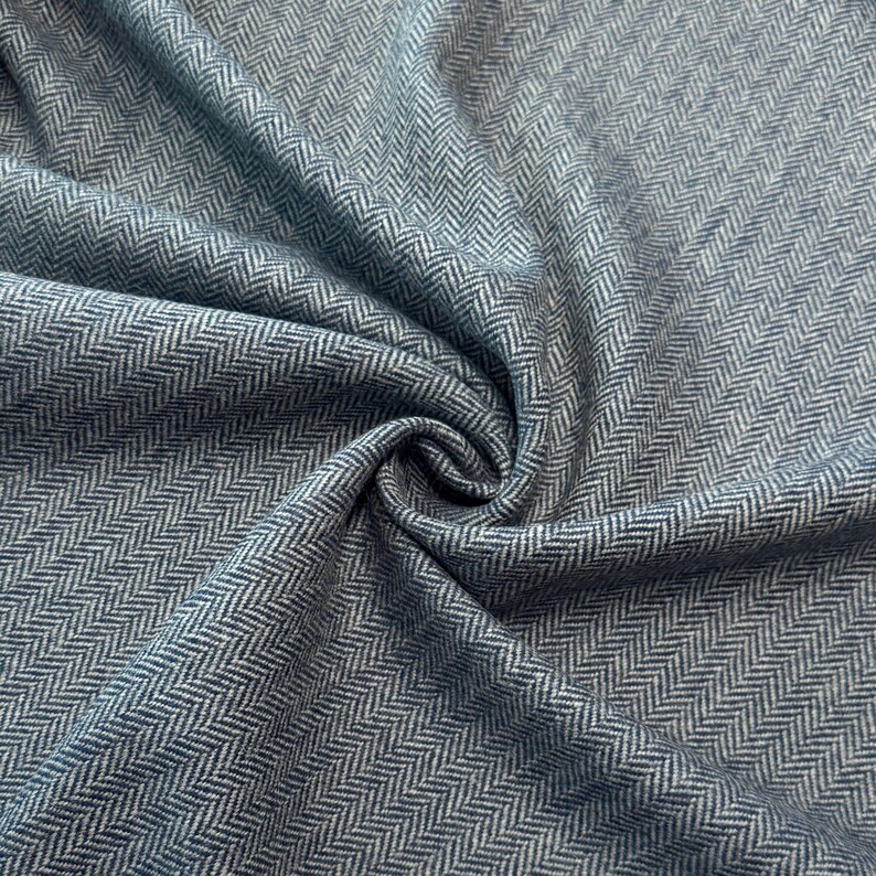 Gefalteter Fischgrät-Tweed in meliertem Blau und Hellgrau, der die hochwertige Schurwollqualität und die moderne Interpretation des klassischen Musters präsentiert. Der Wollstoff ist wärmend und robust, dabei wollig weich und angenehm im Griff.