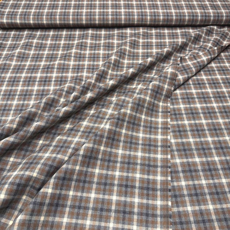 May include: Close-up of a fabric with a brown, white, and navy blue plaid pattern. The fabric is folded and draped, showing the texture and design. The plaid pattern is a classic design.