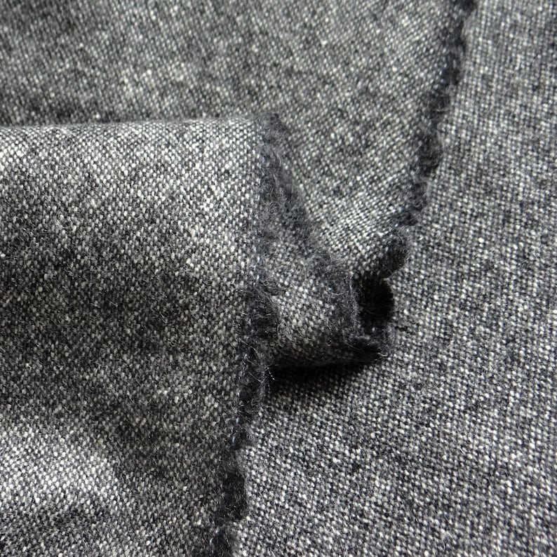 May include: Close-up of a folded piece of gray tweed fabric. The fabric has a textured appearance with a mix of dark gray and white threads. The edges of the fabric are frayed, revealing the weave.