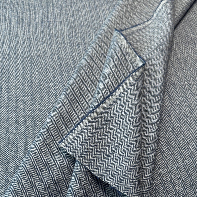 Detailaufnahme eines Tweed-Wollstoffs in Blau und Hellgrau, in Falten gelegt, die die warme, hochwertige Textur, die melierte Farbgebung und das klassische Fischgrätmuster zeigt. Es ist perfekter Stoff für Herbst und Winter. Made in Germany.