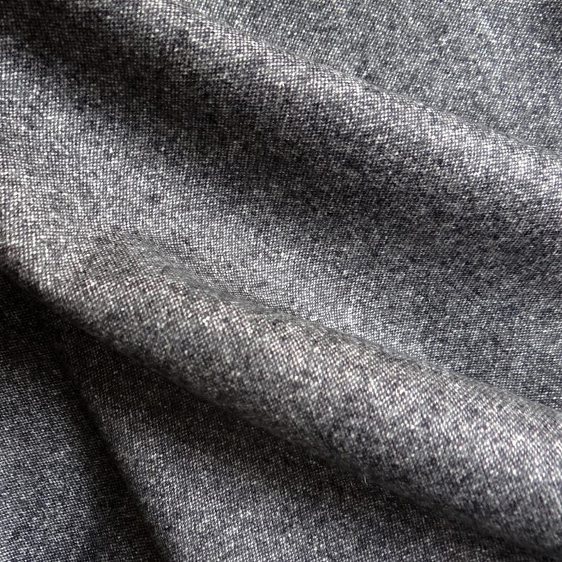 May include: Close-up of a textured gray fabric with subtle variations in tone. The material appears to be woven, with visible threads creating a slightly rough surface. The fabric is draped, showing folds and shadows.